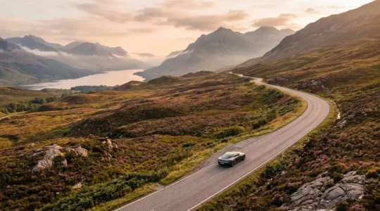 Scenic view of a car traveling through misty Scottish Highlands at dawn with mountains and lochs in background